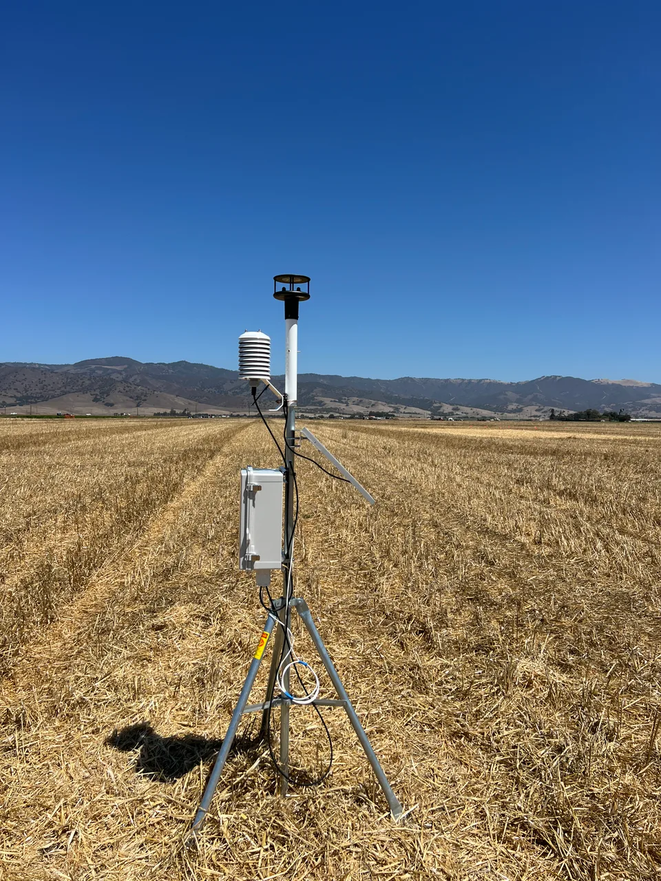 A weather monitoring station on a tripod in a harvested field.